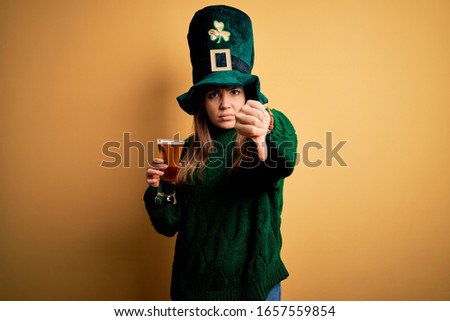Young beautiful woman wearing green hat drinking glass of beer on saint patricks day looking unhappy and angry showing rejection and negative with thumbs down gesture. Bad expression.