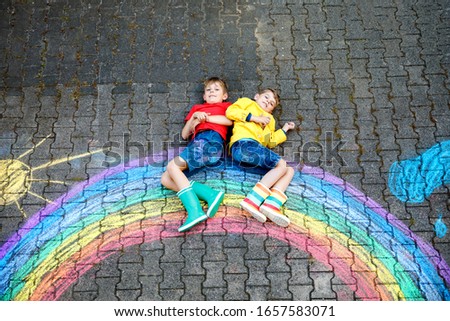 two school kids boys having fun with with rainbow picture drawing with colorful chalks on asphalt. Siblings, twins and best friends in rubber boots painting on ground playing together.