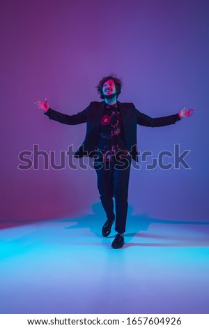 Young caucasian musician in casual dancing on gradient blue-purple background in neon light. Concept of music, hobby, festival. Joyful party host, DJ, stand upper, dancer. Colorful portrait of artist.