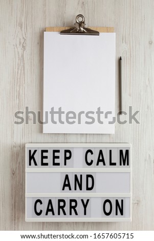 'Keep calm and carry on' words on a lightbox, clipboard with blank sheet of paper on a white wooden surface, top view. Overhead, from above, flat lay. 