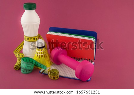 Sport weight loss and fitness concept with a shuttlecock, dumbbell weight and tape measure wound round a white plastic bottle with a slim line shape, open notebook with pen on a bright pink background