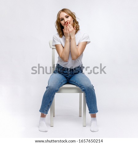 A blonde girl is sitting on a chair and blowing a kiss in a white t-shirt and blue jeans on a white background.
