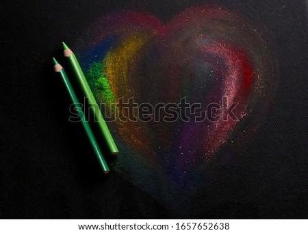 Two green pencils and multicolored painting heart shape are isolated on the black fabric texture background close up taken with copy space around 