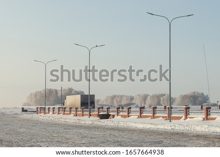 Asphalt road. Trucks in the parking lot. Brick red fence. Concrete poles. Lanterns. Trees and grass in the snow. In the open air.