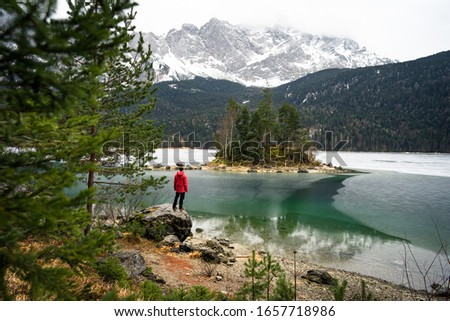 Man in red jacket looking over frozen lake. Little island with trees at lake Eibsee Bavaria Zugspitze. Beautiful winter day 