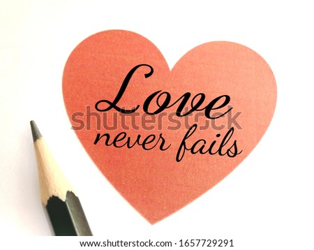 Text on red paper in heart shape saying love never fails and a pencil isolated on white background.