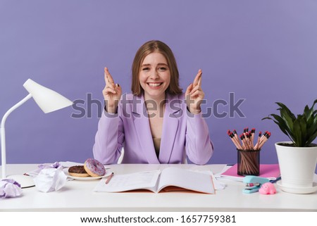 Image of a cheery optimistic young woman sit at the table indoors isolated over purple wall background showing hopeful gesture fingers crossed.