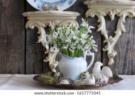 Easter Composition: Fresh flowers, Snowdrops in Porcelain Pitcher, Milk Jug, Creamer, Feathers, Quail eggs on Round Metal Plate, Silver ton Tray, Wall Aged Wooden Scones, Vintage Style, Daylight 