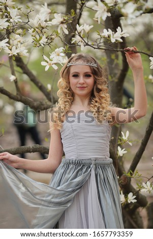 
young beautiful blonde girl in a silver dress with a train in the spring garden where white magnolia blooms