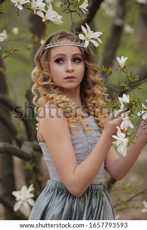 
young beautiful blonde girl in a silver dress with a train in the spring garden where white magnolia blooms