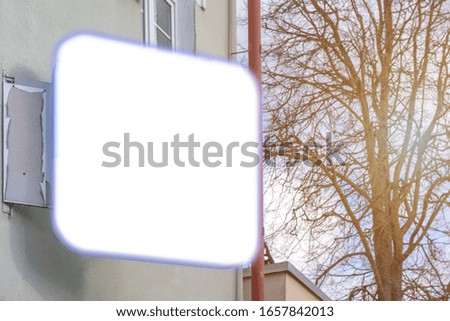 white blank sign with space for poster on building wall in city street against bare tree at bright sunlight on spring day