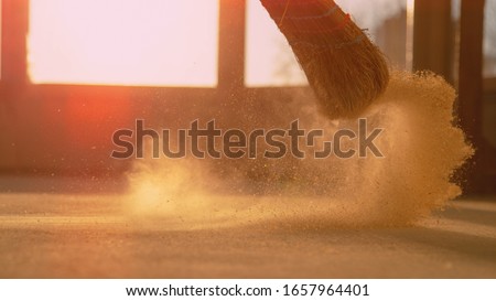 LENS FLARE, CLOSE UP, LOW ANGLE: Dust gets swept up into air as an unrecognizable person cleans the construction site floor. Contractor sweeps the ground of a construction site with a straw broom Royalty-Free Stock Photo #1657964401
