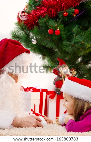 Close up of old Santa Clause giving small present to little girl. Laying together under Christmas tree 