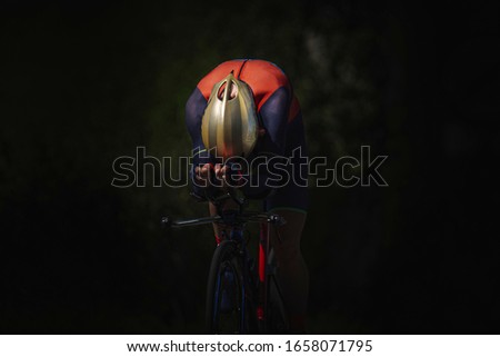 Unknown male cyclist riding on aerodynamic mode in the dark. He is using a time trial bike. He is wearing gold helmet and red and blue cycling gear. He is alone. Royalty-Free Stock Photo #1658071795