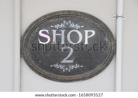 A rock-textured oval sign against a white wall reading "Shop 2" in reflective metal and with decorative patterns above and below