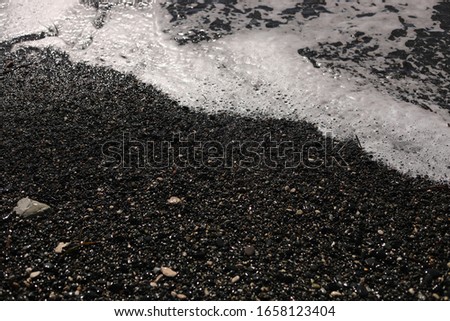 Shallow volcanic black sandy beach and pebbles, water waves and white foam washes the coastline with wet dark sand on the sea coast. Ocean shore natural background with copy space. selective focus
