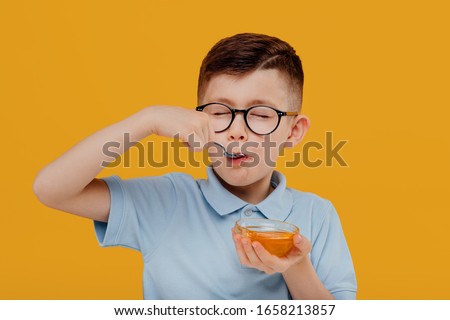 Happy little kid boy in glasses smiling and eating fresh liquid honey from bowl against yellow background Royalty-Free Stock Photo #1658213857