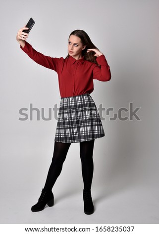 full length portrait of a pretty brunette girl wearing a red shirt and plaid skirt with leggings and boots. Standing pose, holding a phone, against a studio background.
