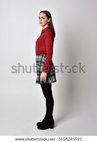full length portrait of a pretty brunette girl wearing a red shirt and plaid skirt with leggings and boots. Standing pose in side profile against a studio background.
