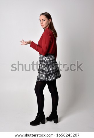 full length portrait of a pretty brunette girl wearing a red shirt and plaid skirt with leggings and boots. Standing pose in side profile against a studio background.
