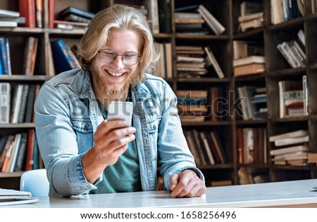 Young bearded man student entrepreneur hipster teacher glasses office home library desk hold modern smart phone use app messenger read text message watch video call smile laugh at screen copy space. Royalty-Free Stock Photo #1658256496