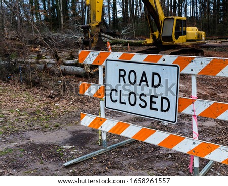 Road closed sign attached to orange and white striped, plastic barriers on metal frame, at road construction site. A backhoe is clearing fallen trees in the background
