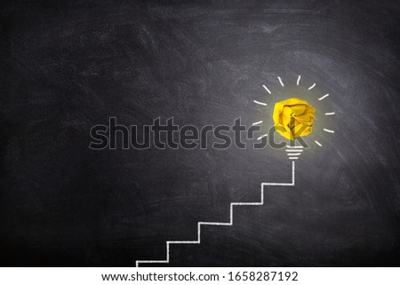 Business Idea and Success Concept :  Yellow crumpled paper ball light bulb lighting grow on the top of white chalk ladder handwriting on chalkboard.