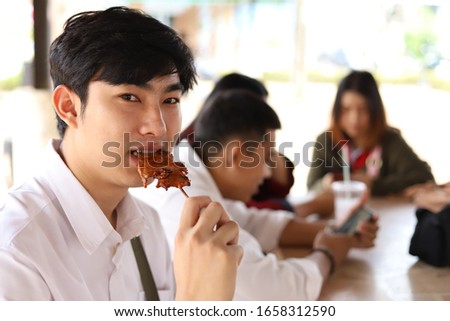 Teen boy eating grilled pork or barbecue with friends while having socializing activity in the university campus, selective focused picture of a lad taking a bite on tasty meat, lifestyle concept pic