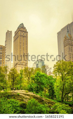 View of large bolder in Central Park, New York, surrounded with trees; people taking photos from the rocks; New York skyline in background