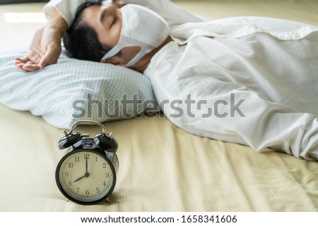 Asian businessman wearing surgical mask sleeping on the bed with alarm clock
