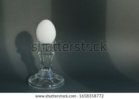 A white chicken egg in a vintage classic glass candlestick holder on a textured gray background with a shadow. Creative minimal modern concept of food, Easter.