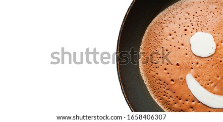 Banner with white space for text. A pancake made of liquid chocolate dough with bubbles is fried in a black pan with drops of fat and oil on an induction cooker. Funny food with a face image.