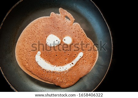 A pancake made of liquid chocolate dough with bubbles is fried in a black pan with drops of fat and oil on an induction cooker. Funny food with a face image.