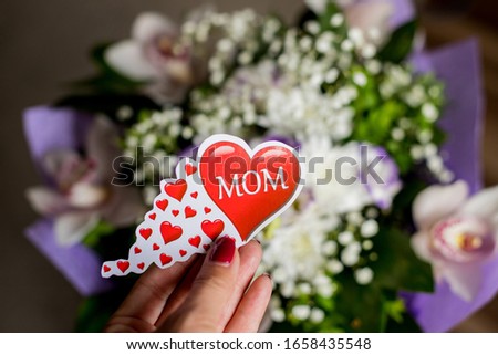 Bouquet of orchids, Lisianthus,Gypsophila.Female hand is holding a big red heart with text mom, Present with love.MOTHERS DAY. womens day or happy birthday gift