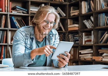 Young bearded man student entrepreneur hipster teacher glasses office home library desk hold modern tablet computer use app surf net social network watch video call smile tap touch screen copy space. Royalty-Free Stock Photo #1658487397