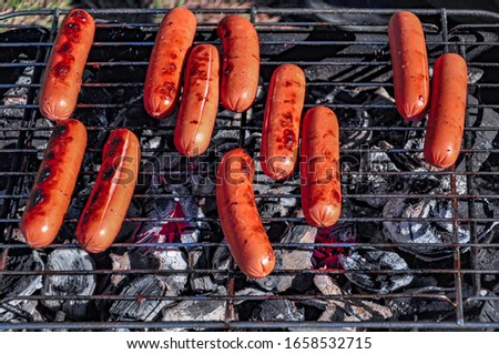 Standing on the grass, a small portable picnic grill with burning coals and sausages roasting on the grill, brightly lit by the rays of the setting sun