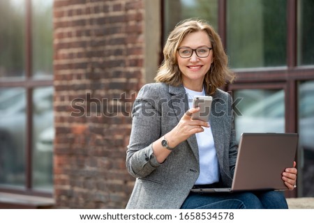 Portrait of a happy mature business woman using her mobile phone and laptop, outdoors, near the office. Royalty-Free Stock Photo #1658534578