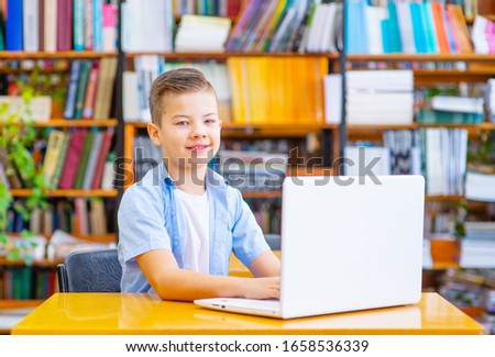 Boy sitting at the table in the library at the laptop, looking at the camera with a smile