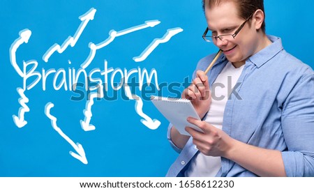 Man in casual clothes with glasses on a blue background sits on a chair holding a notebook and nibbling on a pen while doing a brainstorm session. Abstract inscription with outgoing arrows. Close up.