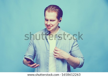 A man in casual clothes on a blue background controls a playlist in a smartphone while listening to music on wireless headphones. Toned. Close up.