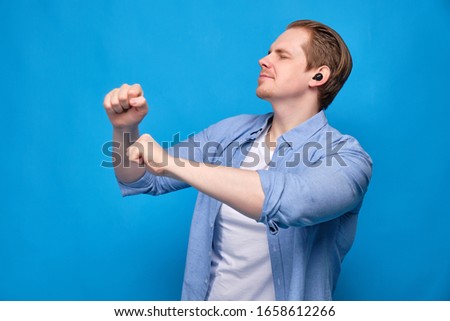 A man in casual clothes on a blue background with his eyes closed listens to music in wireless headphones and dances. Close up.