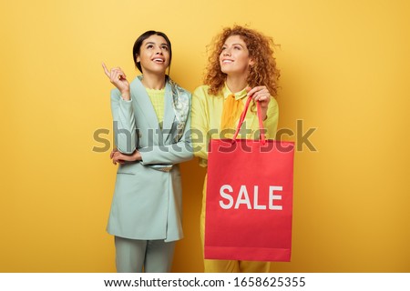 happy redhead girl holding shopping bag with sale lettering near smiling african american woman pointing with finger on yellow