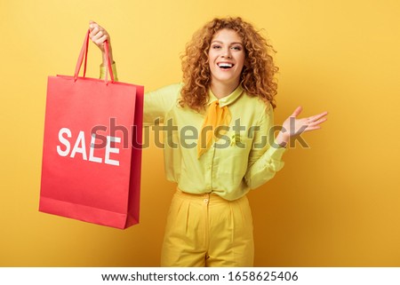 cheerful redhead woman holding shopping bag with sale lettering on yellow