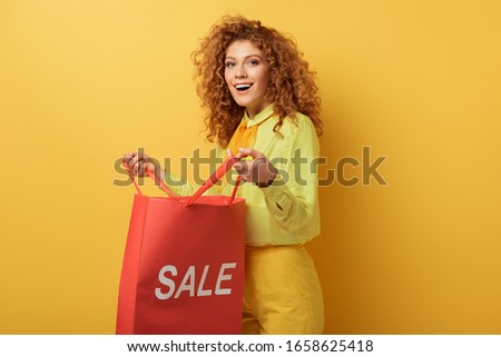 happy redhead woman holding shopping bag with sale lettering on yellow