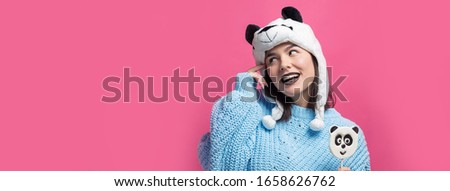 Funny young girl standing with yummy panda-lollipop in her hand and a hat on his head on a pink background.