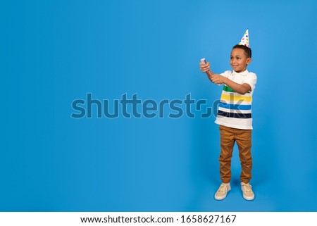 African American boy blows up flapper holding her in his hand dressed in white polo and party cap stands on blue studio background. Birthday concept and holidays party concept with place for text.