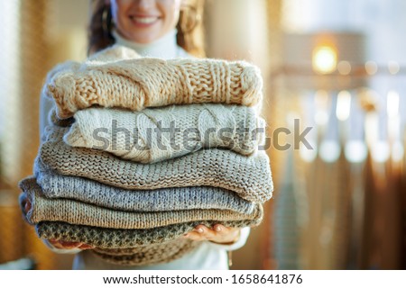 Closeup on happy elegant middle age housewife in white sweater and skirt at modern home in sunny winter day holding pile of sweaters.