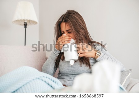 Sick young woman sitting on sofa blowing her nose at home in the sitting room. Photo of sneezing woman in paper tissue. Picture showing woman sneezing on tissue on couch in the living-room
