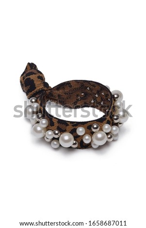 Subject shot of a spotted fabric scrunchy tied in a knot and adorned with lots of silver and nacre pearls. The hair band with animalistic print is isolated on the white background. 