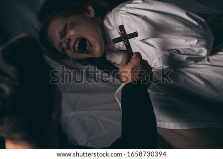 exorcist holding cross over demonic yelling girl in bed Royalty-Free Stock Photo #1658730394